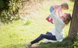 Kobieta w ciąży siedząca przy drzewie i trzymająca w górze dziecko