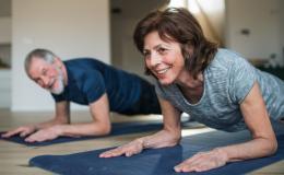 Starsza para ćwiczy w domu, twarzą do podłogi opierają się łokciami na macie