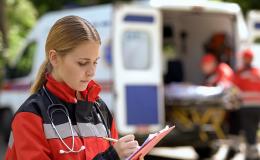 Na pierwszy planie kobieta zapisująca informacje w notesie. Na drugim planie karetka, z której 2 osoby wyjmują nosze.