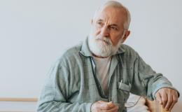 Starszy mężczyzna z siwymi włosami i brodą siedzi na łóżku i patrzy w dal.