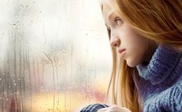 Smutna dziewcynka z długimi blond włosami patrzy przez szybę w dal. Za oknem pada deszcz.