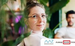 Odwrócona w stroną oglądającego siedzaca kobieta w okularach. Na drugim placie siedzący mężczyzna