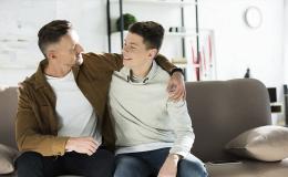 Ojciec obejmuje ramieniem nastoletniego syna, siedzą na kanapie