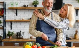 Kobieta i mężczyzna w kuchni. Mężczyzna kroi warzywa, kobieta obejmuje go za szyję.