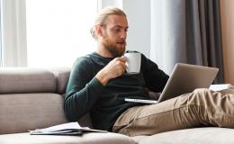 Brodaty młody facet z kubkiem na kanapie i laptopem na kolanach