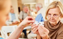 Starsza kobieta w okularach trzyma dwa puzzle i próbuje je dopaswać. Ktoś jej pomaga.