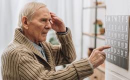 Starszy mężczyzna patrzy na kalendarz i wskazuje datę. Drugą ręką dotyka skroni. Wygląda na zdezorienrowanego.