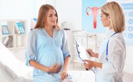 Młoda, uśmiechnięta kobieta w ciąży siedzi na kozetce i trzyma się za brzuch podczas rozmowy z lekarką.