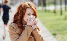 Kobieta, która jest uczulona na pyłki, kicha i zasłania nos chusteczką. Stoi na chodniku, ma rude włosy.