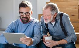 Dorosły syn z ojcem siedzą na schodach w domu. Syn pokazuje coś ojcu na tablecie, senior ma w dłoniach kubek.