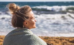 Młoda kobieta w blod włosach spiętych w koczek siedzi na plaży. Patrzy w stronę morza, słońce oświetla jaj twarz. Na ramionach ma koc.