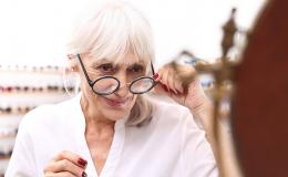 Starsza pani przymierza okulary u optyka. Ma siwe włosy i czerwone paznokcie.