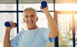 Starszy mężczyzna ćwiczy. Podnosi ciężarki na wysokość głowy. Ma siwe włosy, błękitny t-shirt. Uśmiecha się.