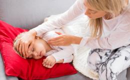 Matka trzyma dłoń na czole córki, sprawdza jej tempreraturę. Dziewczynka leży pod kocem.