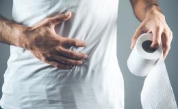 Mężczyzna w białej koszulce. Zbliżenie na jego bolący brzuch, na którym trzyma rękę. W drugiej ma rolkę papieur toaletowego.
