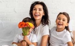 Małoda matka z dwójką kilkuletnich dzieci siedzi na kanapie. Wszyscy się uśmiechają. Chłopiec zakrywa twarz bukietem pomarńczowych kwiatów.