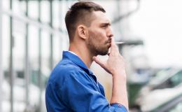 Profil mężczyzny, który stoi przed warsztatem i pali papierosa. Jest ubrany w kombinezon, ma lekki zarost.