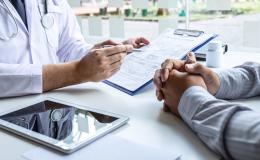 Lekarz omawia wyniki z pacjentem. Pokazuje mu jakiś wydruk. Zbliżenie na ręce na biurku.