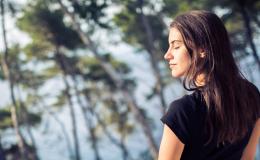 Kobieta stoi nad morzem, w tle rosną sosny. Ma zamknięte oczy, twarz wystawia do słońca. Delektuje się chwilą