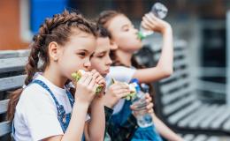 Trójka dzieci siedzi na ławce przed szkołą i je drugie śniadanie. Na pierwszym planie dziewczyna z kanapką z której wystaje sałata, w tle inna pije wodę.
