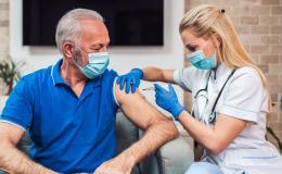 Straszy mężczyna w maseczce siedzący na fotelu. Młoda bobieta w białym fartuchu podaje mu szczepionkę w ramię.