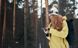 Dziewczyna w żółtej kurtce i czapce na tle lasu. Sprawdza coś w telefonie.