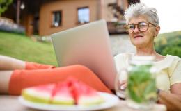 Siwa pani z laptopem przed domem w górach, laptop na kolanach, nogi na stole, na stole arbuz, w tle dom na stoku