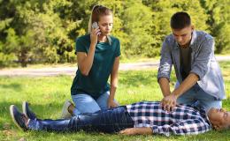 park, poszkodowany leży, dziewczyna dzwoni, chłopak robi masaż serca