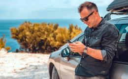 Mężczyzna oparty o auto, z telefonem, w tle krajobraz środziemnomorski