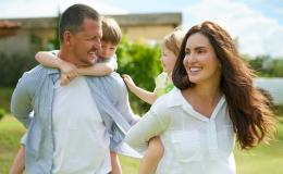 młodzi rodzice niosą na barana swoje dzieci, w tle dom na wsi