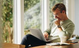 mężczyzna siedzi przy oknie balkonowym z laptopem na kolanach