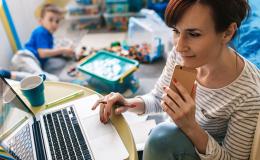 Mama przed komputerem, w jednej ręce komórka, w tle na podłodze bawi się chłopczyk