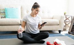 dziewczyna w bałym t-skircie siedzi po turecku w domu, ma w ręku butelkę wody, w drugiej komórkę