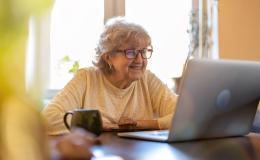 starsza pani w swetrze przy stole przed laptopem i kubkiem herbaty