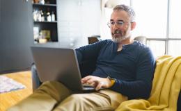 Pan z siwą brodą na kanapie z laptopem na kolanach