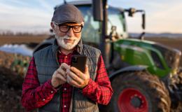 Pan z siwą brodą z komórką, w tle traktor