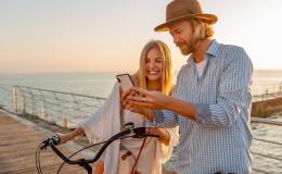 Dziewczyna i chłopak stoją na molo . Dziewczyna trzyma oburącz kierownicę roweru. Mężczyzna pokazuje jej coś na telefonie. W tle morze.