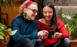 Dwie kobiety siedzą na schodach wśród zieleni i sprawdzają coś na telefonie.