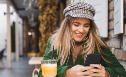 diewczyna w kyrtce i czapce z daszkiem pije kawę przy stoliku na ulicy i patrzy w komórkę