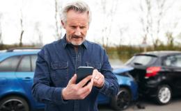 w tle zderzenie 2 aut mężczyzna z siwą bródką patrzy na telefon