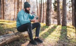 brodaty mężczyzna w lesie z butelką wody,siedzi na pniu i patrzy na komórkę