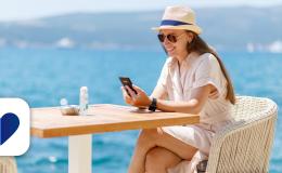 Kobieta siedzi prrzy stoliku, na stole butelka wody, ona w kapeluszu i ciemnych okularach, z komórką, w tle morze