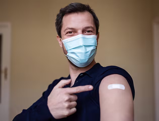 młody mężczyzna w maseczce pokazuje na ramię z plastrem po szczepieniu