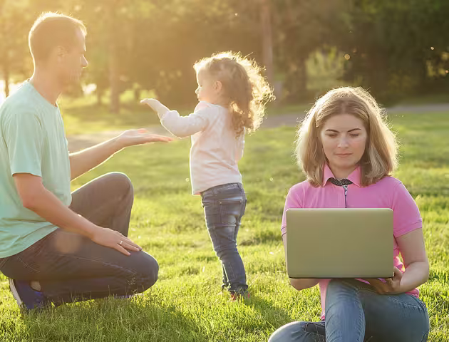 Ojciec bawi się z córeczką na łące, matka zagląda do laptopa