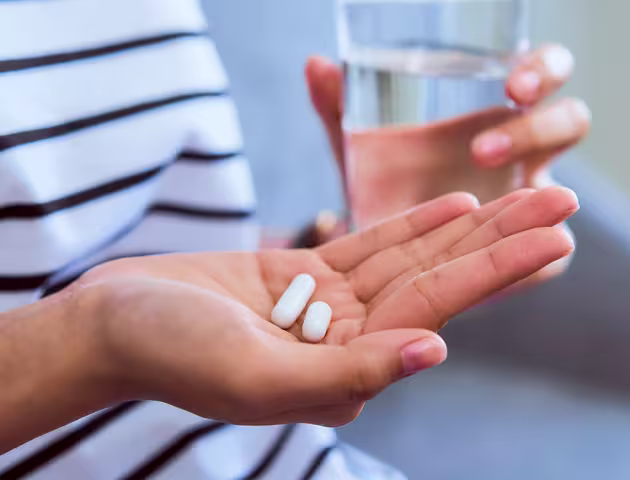 Wykadrowane ręce siedzącej osoby. W prawej ręce 2 tabletki, a w lewej szklanka z wodą