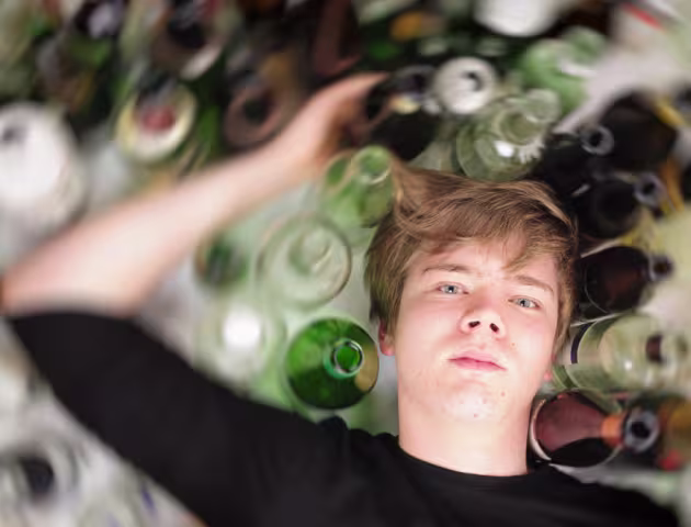 Nastolatek leży na podłodze, a dookoła niego stoją puste butelki po alkoholu.