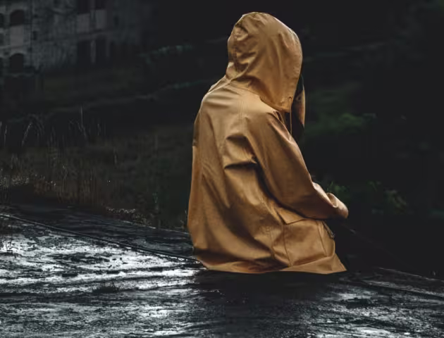 Siedząca tyłem na zaniedbanym wyłomie budynku osoba w żółtym płaszczu przeciwdeszczowym, z kapturem na głowie