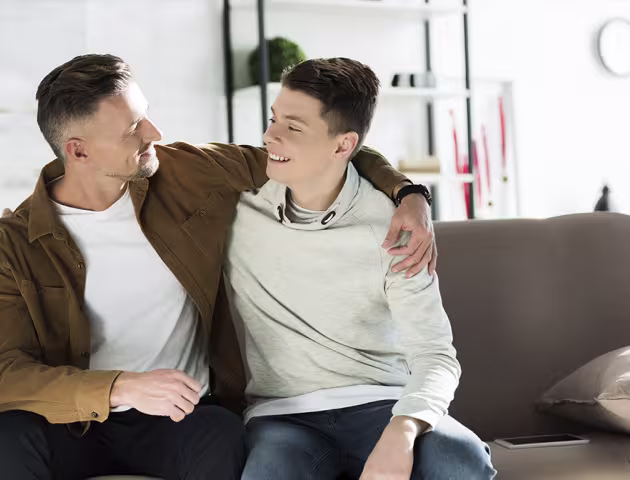 Ojciec obejmuje ramieniem nastoletniego syna, siedzą na kanapie