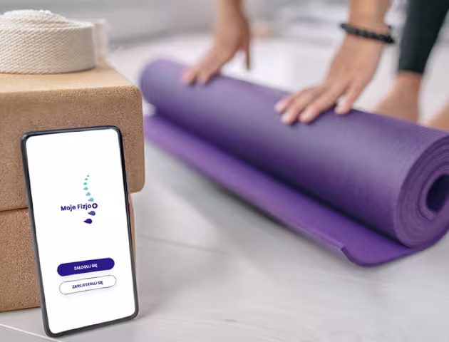 Smartfon z aplikacją Moje Fizjo na ekranie. W tle ręce rolujące matę do ćwiczeń.