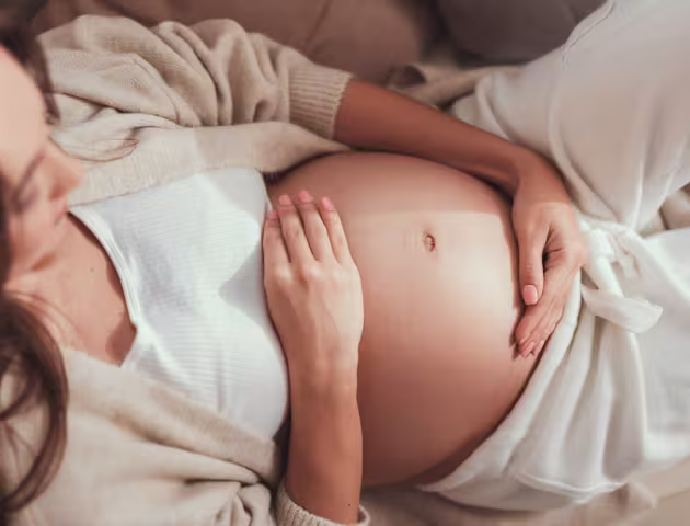 Leżąca kobieta w ciąży obejmuje brzuch dłońmi.